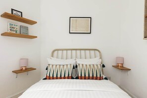 A bedroom with a bed, two bedside lamps, a framed picture above the bed, and a shelf with books and a framed picture. - New! Cosy 3BR w/Patio | Shoreditch & Liverpool St (Greater London)