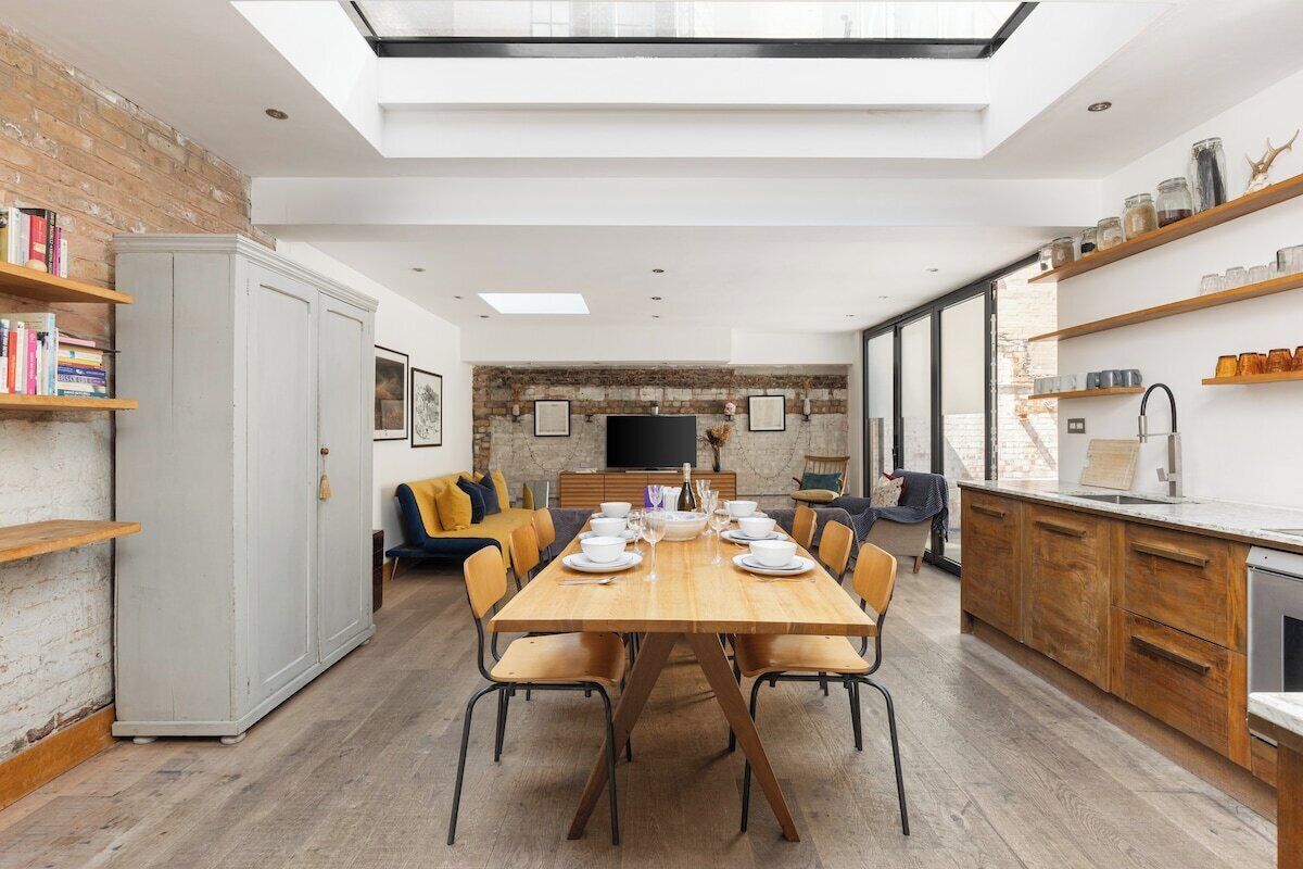 A modern kitchen with a dining area, featuring a wooden table set for four, a built-in oven, and a brick wall.