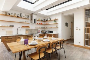 A modern kitchen with wooden shelves, a dining table set for six, and a skylight above. - New! Cosy 3BR w/Patio | Shoreditch & Liverpool St (Greater London)