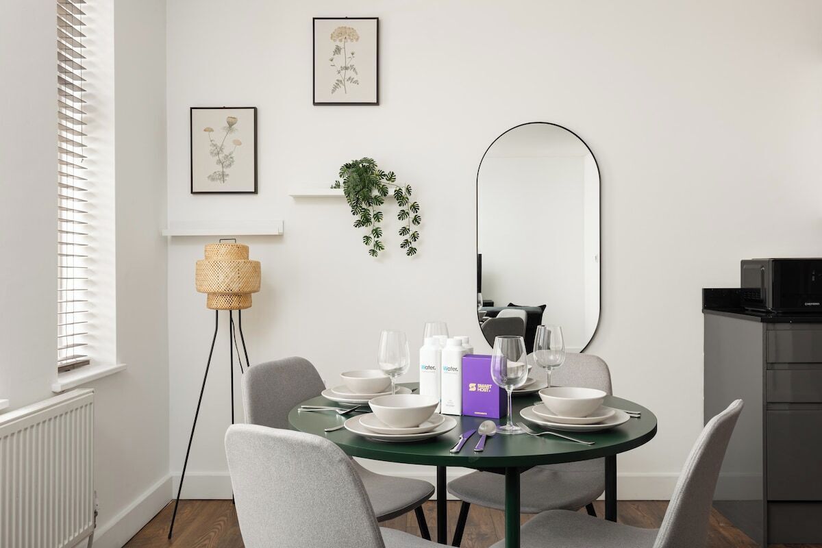 A dining area with a round table set for two, a mirror, framed botanical prints, and a potted plant.