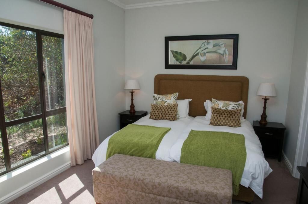 A bedroom with a large window, a bed with green and white bedding, two bedside tables with lamps, and a framed picture on the wall.