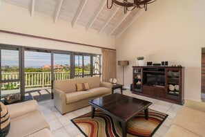 A spacious living room with a sofa, coffee table, and entertainment unit, featuring large windows and a balcony.