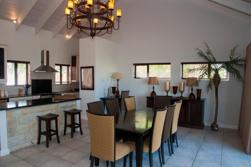 A dining area with a central table, chairs, a chandelier, and a kitchen in the background.