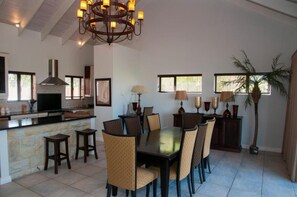 A dining area with a central table, chairs, a chandelier, and a kitchen in the background.