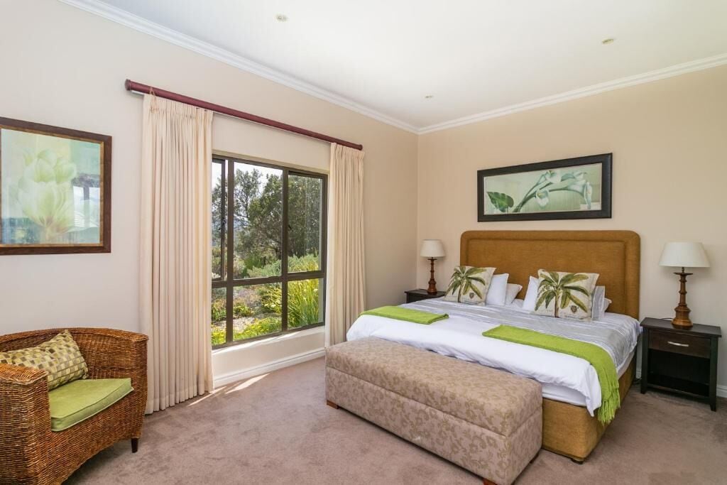 A bedroom with a large window, a bed with a headboard, a nightstand, a lamp, a sofa, and a painting on the wall.