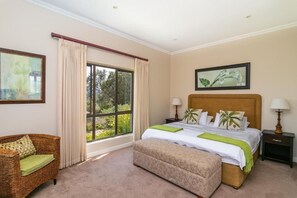 A bedroom with a large window, a bed with a headboard, a nightstand, a lamp, a sofa, and a painting on the wall.