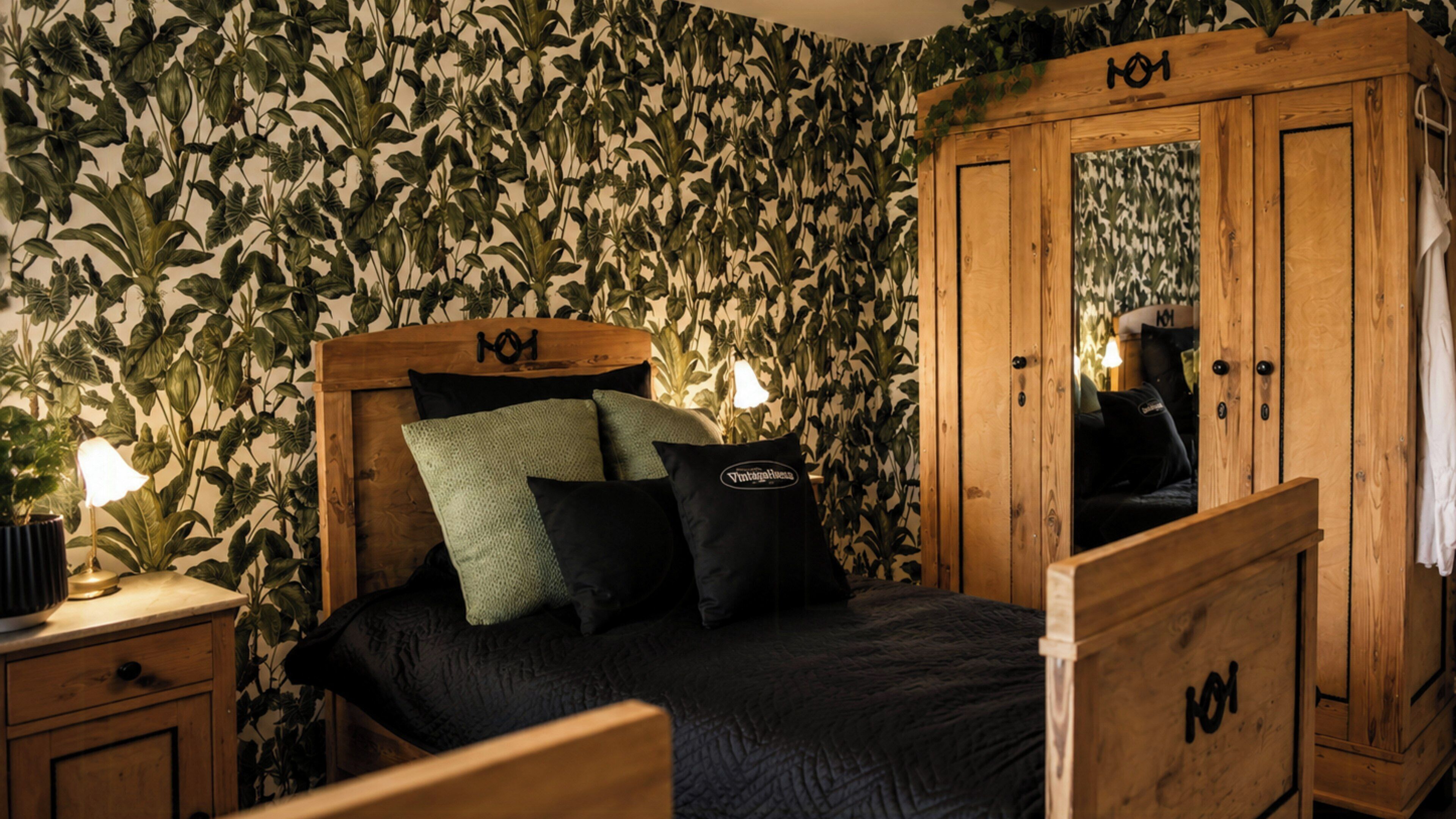 A bedroom with a bed, a wooden wardrobe, and a wall with a leafy pattern.