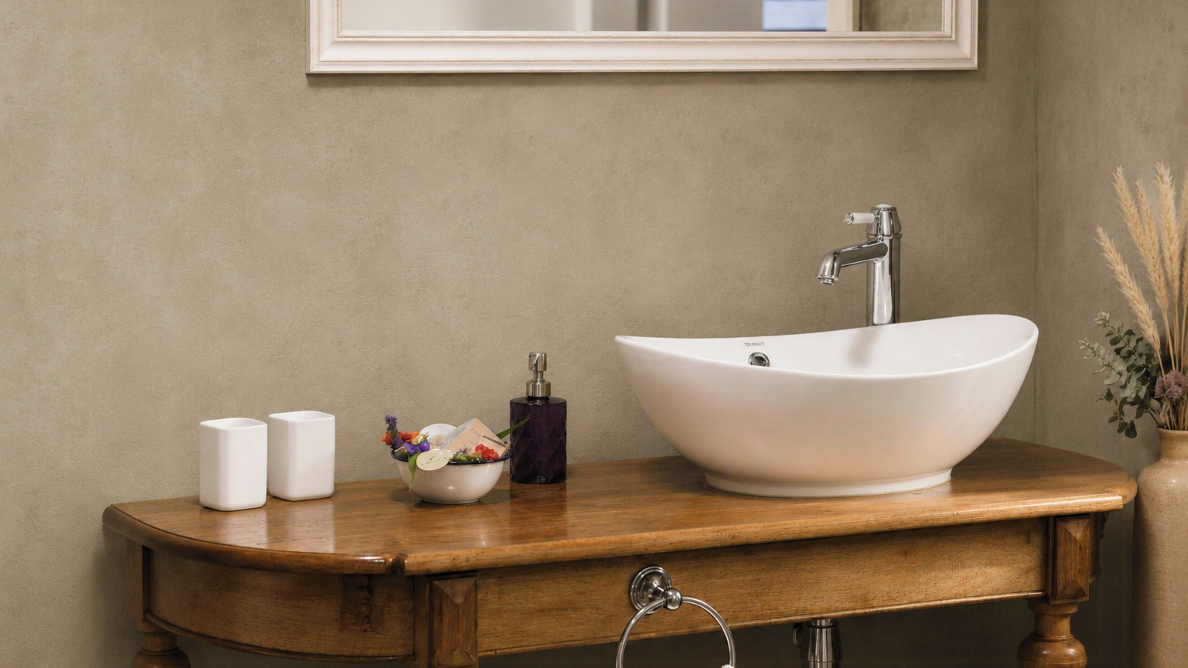 A bathroom with a large white vessel sink, a wooden vanity, two white candles, a small bowl with flowers, and a vase with dried plants.