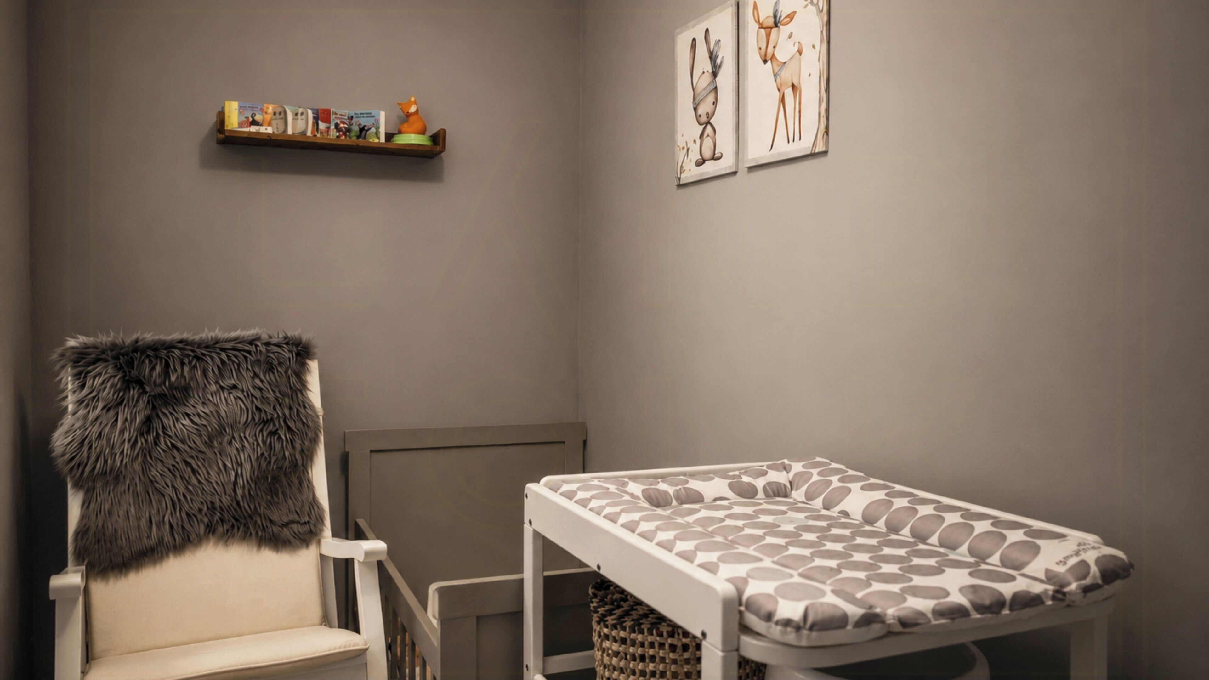 A room with a fur-covered chair, a crib with a patterned mattress, a shelf with books and a toy, and two framed animal illustrations on the wall.