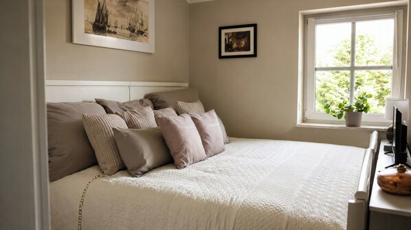 A bedroom with a bed, pillows, a framed picture, a window with a plant, and a painting on the wall.