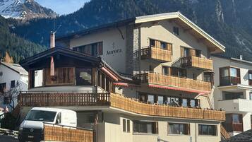 Un hotel de varios pisos con balcones, un letrero que dice 'Aurora' y un paisaje montañoso de fondo.
