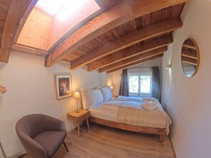 A bedroom with a wooden ceiling, a bed, a chair, a bedside table with a lamp, and a window with curtains. - Apartment  by Interhome (Lenzerheide)
