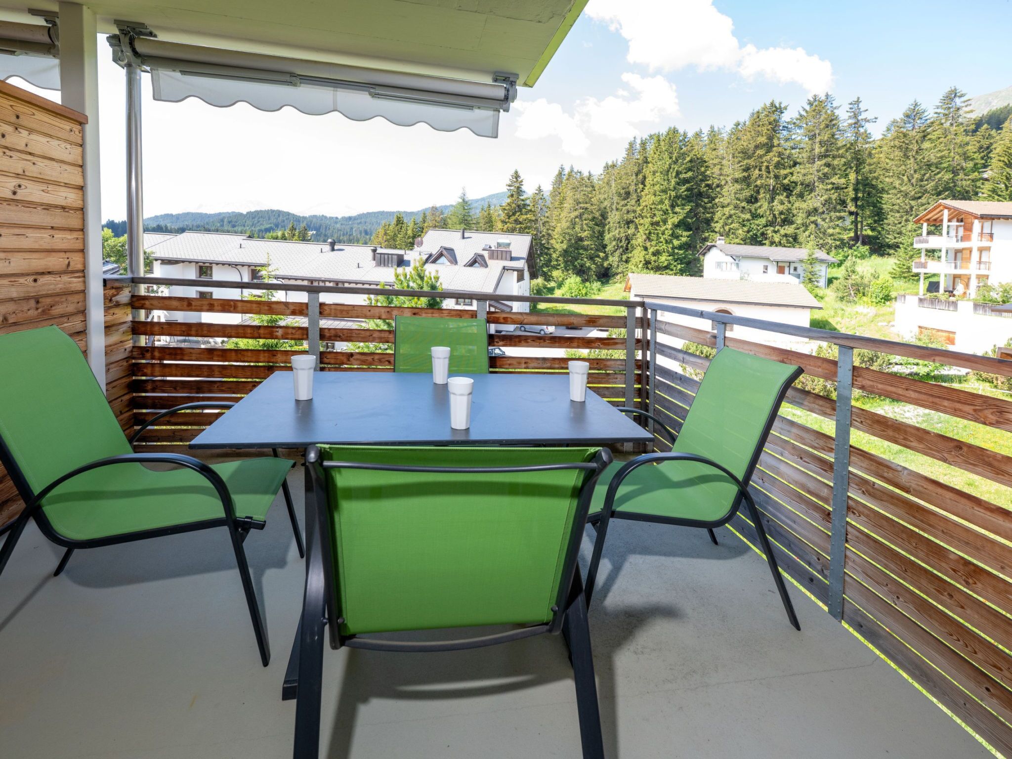Un balcón con mesa y sillas, con vista a una zona boscosa con casas.