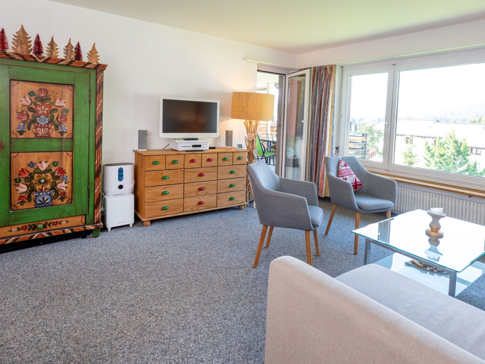 Ein modernes Wohnzimmer mit einem Flachbildfernseher, einem Holzschrank mit bunten Griffen, zwei grauen Sesseln und einem Glastisch.