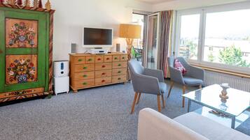 Ein modernes Wohnzimmer mit einem Flachbildfernseher, einem Holzschrank mit bunten Griffen, zwei grauen Sesseln und einem Glastisch.