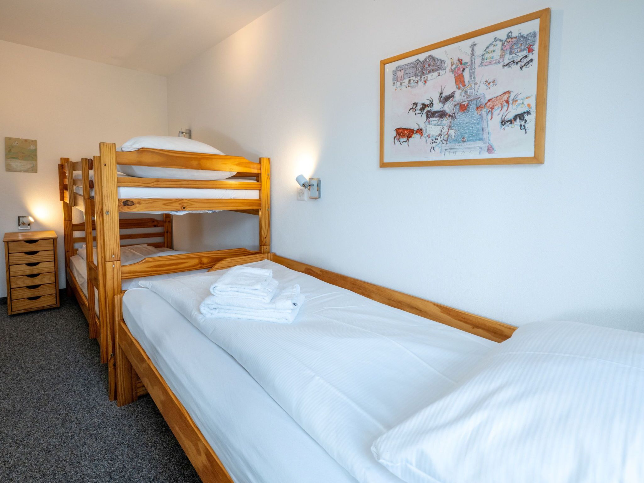 Una habitación con literas, una cama individual, un pequeño aparador de madera y una lámpara fijada a la pared.