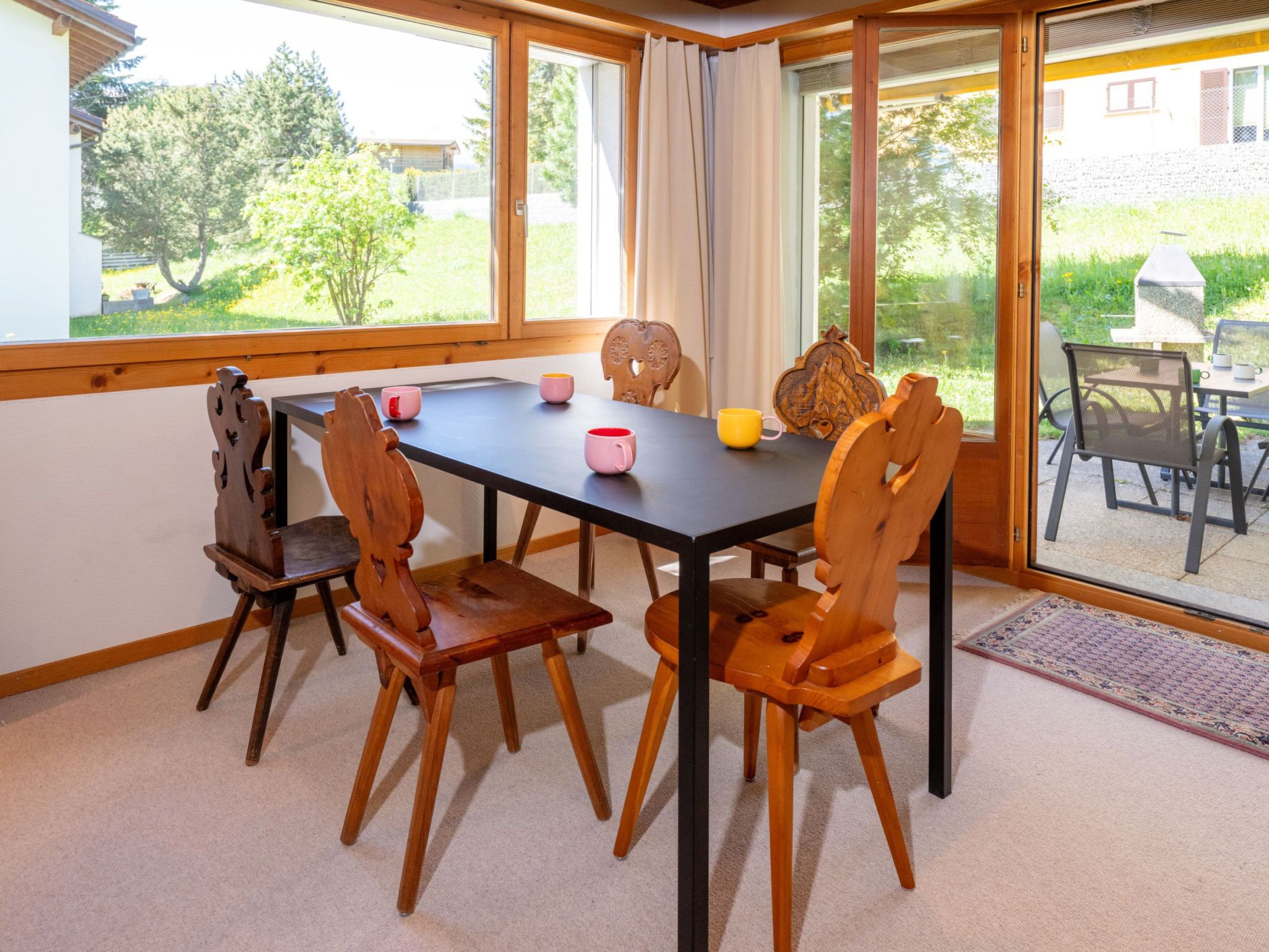 Ein Essbereich mit Tisch und Stühlen, Blick ins Freie durch große Fenster und eine Terrasse mit Grill.