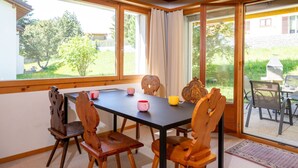 En matplats med ett bord och stolar, utsikt över omgivningarna genom stora fönster och en uteplats med en grill.