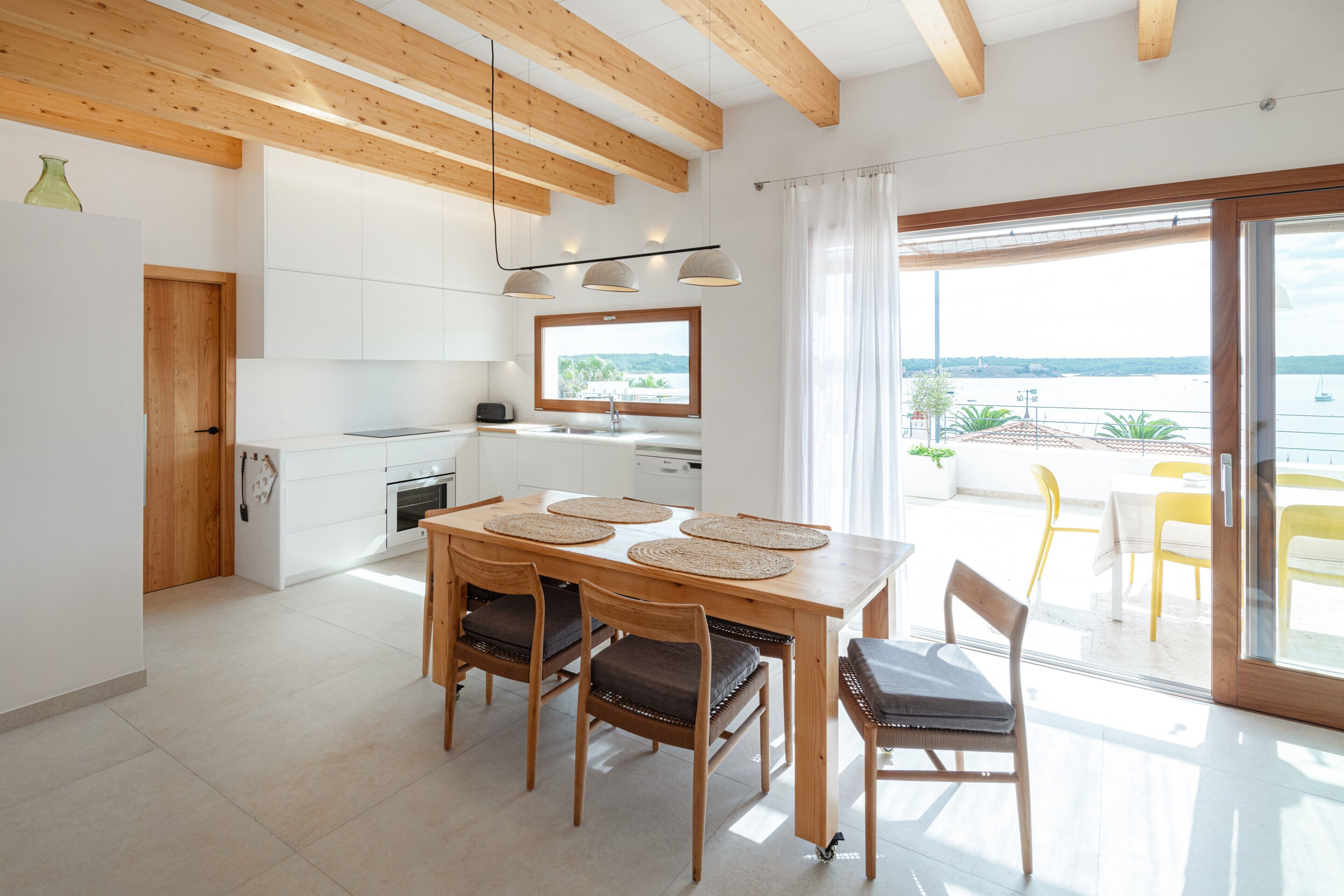A modern kitchen with white cabinets, a wooden dining table with four chairs, and a view of the sea through a large window.
