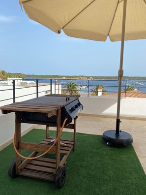 A wooden barbecue grill with wheels on artificial grass, under a beige umbrella, overlooking a marina with boats. - Villa in the exclusive Fornells area with beautiful ocean views. Private Pool (Fornells)