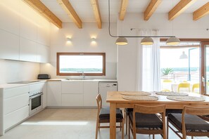 A modern kitchen with white cabinets, a wooden dining table with chairs, and a view of the sea through a large window. - Villa in the exclusive Fornells area with beautiful ocean views. Private Pool (Fornells)