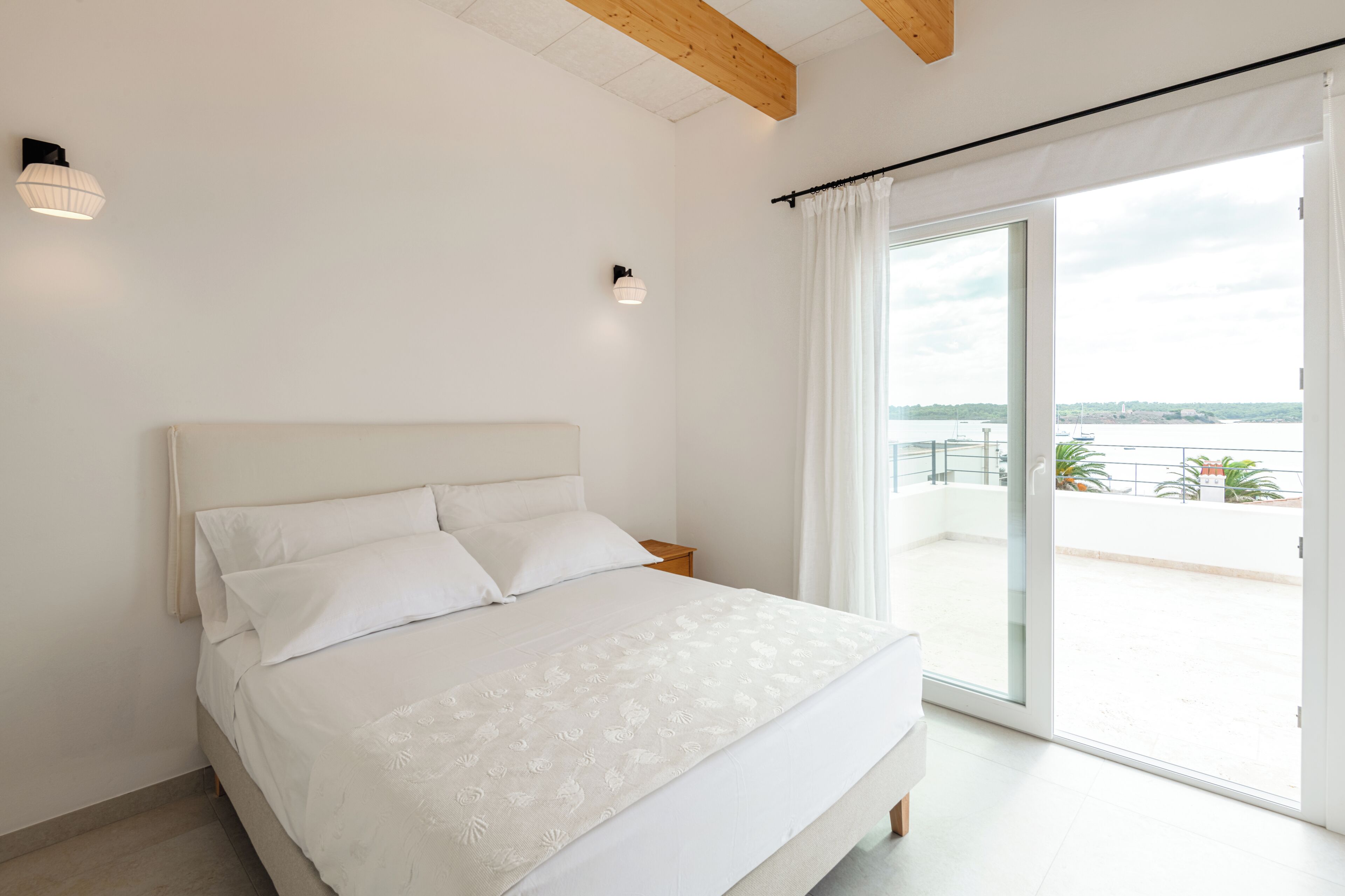A bedroom with a bed, a nightstand, a window with a view of the sea, and a ceiling with exposed beams.