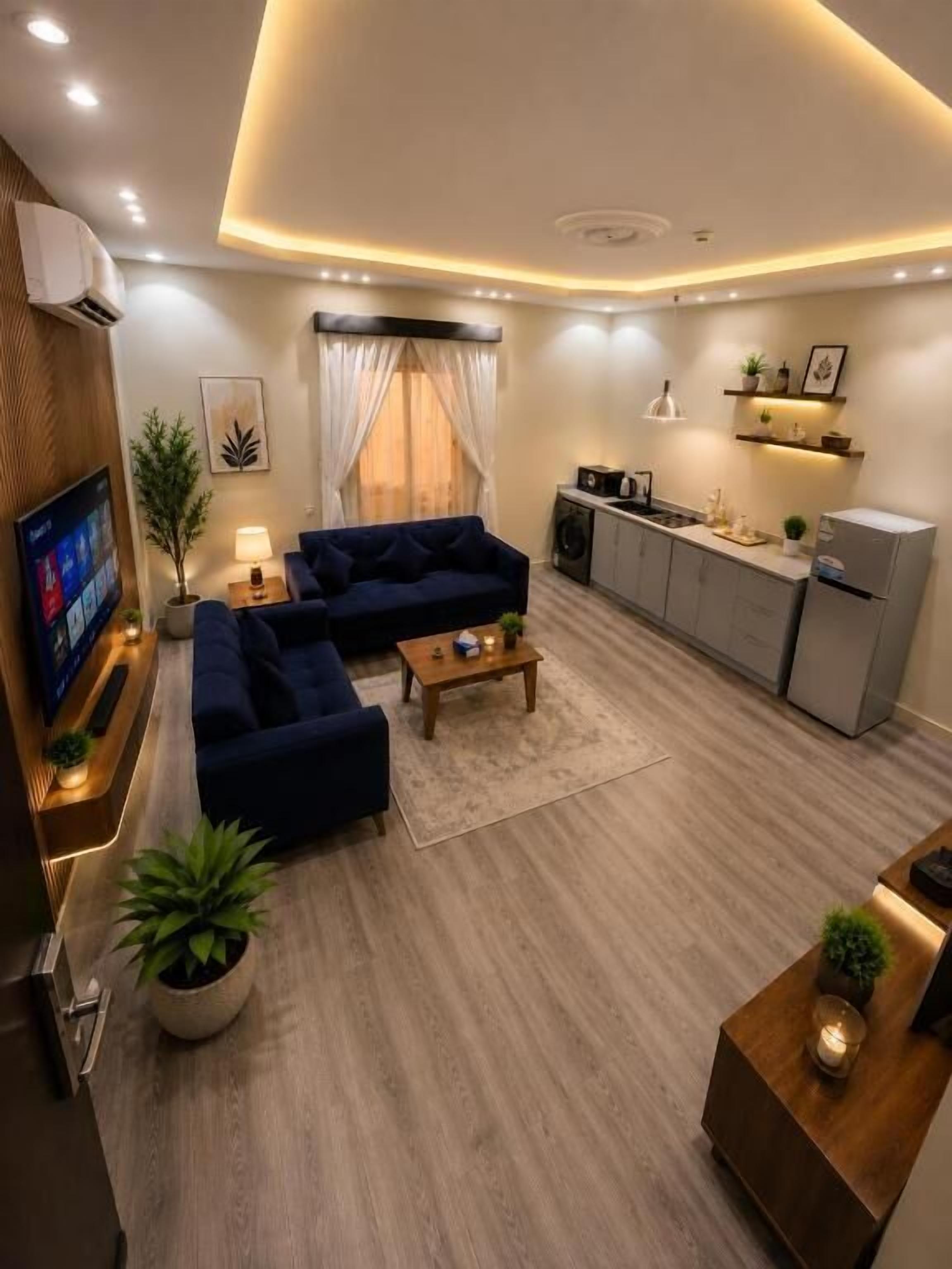 A modern living room with a sofa, coffee table, and a TV. There is a kitchen area with cabinets and a refrigerator.