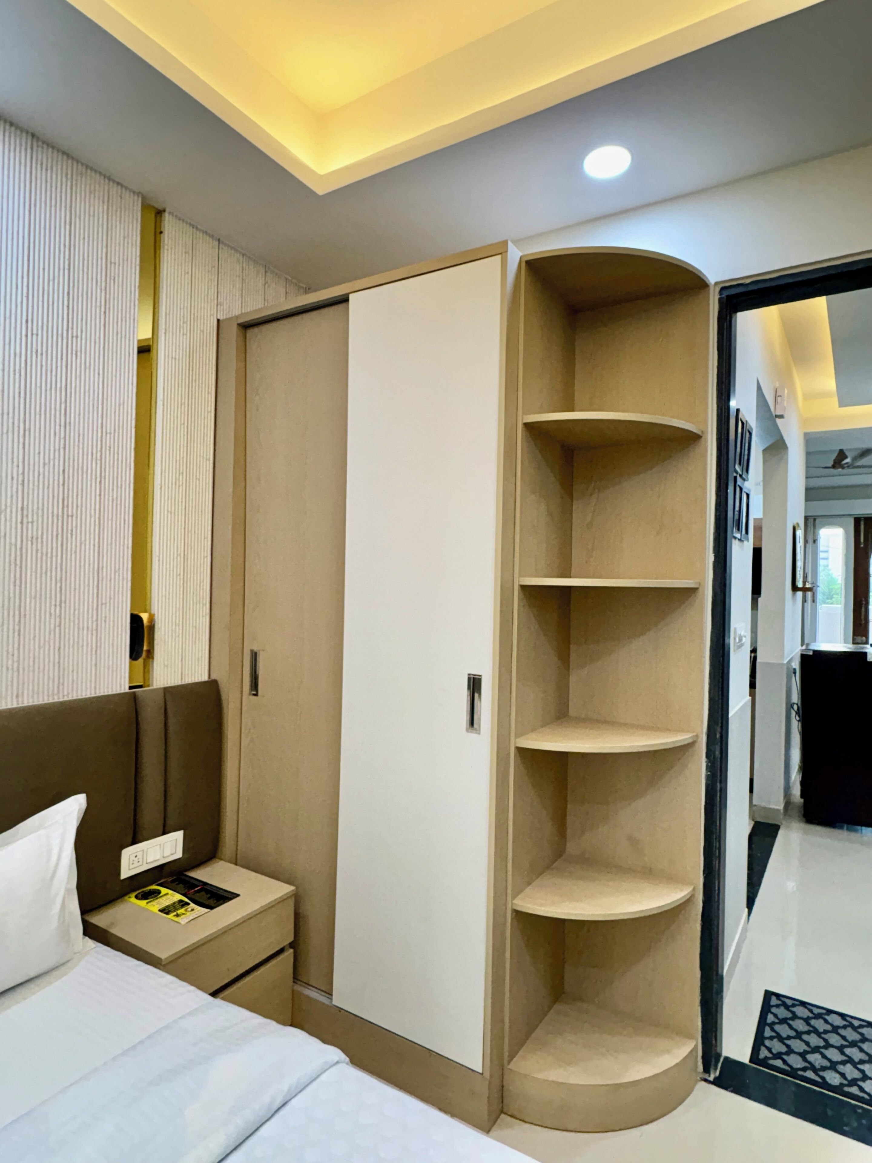 A hotel room with a bed, a wardrobe with shelves, a bedside table, and a nightstand.