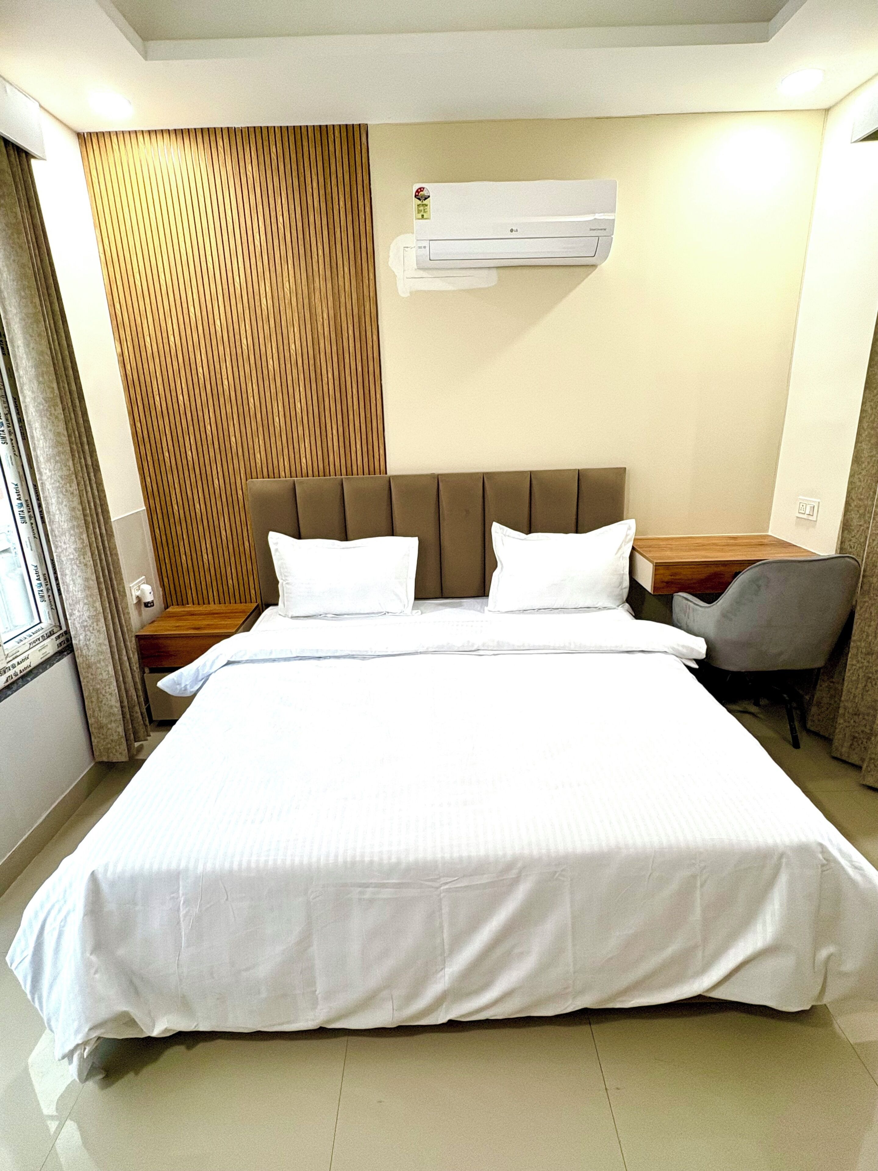 A hotel room with a large bed, a wooden panel wall, an air conditioning unit, a desk with a chair, and a window with curtains.