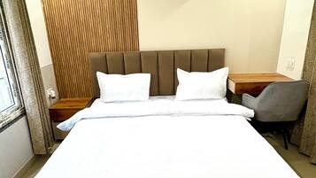 A hotel room with a large bed, a wooden panel wall, an air conditioning unit, a desk with a chair, and a window with curtains.