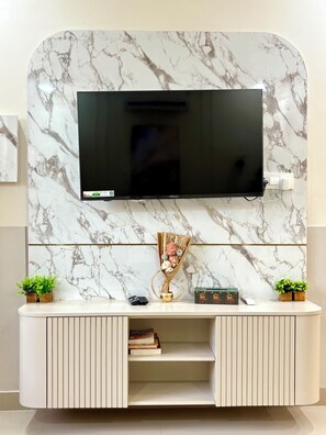 A flat-screen TV mounted on a marble-patterned wall above a white console with a shelf, two remote controls, and a decorative vase. - 3BHK Villa Madhav Nikunj By Iraaya M1 (Vrindavan)