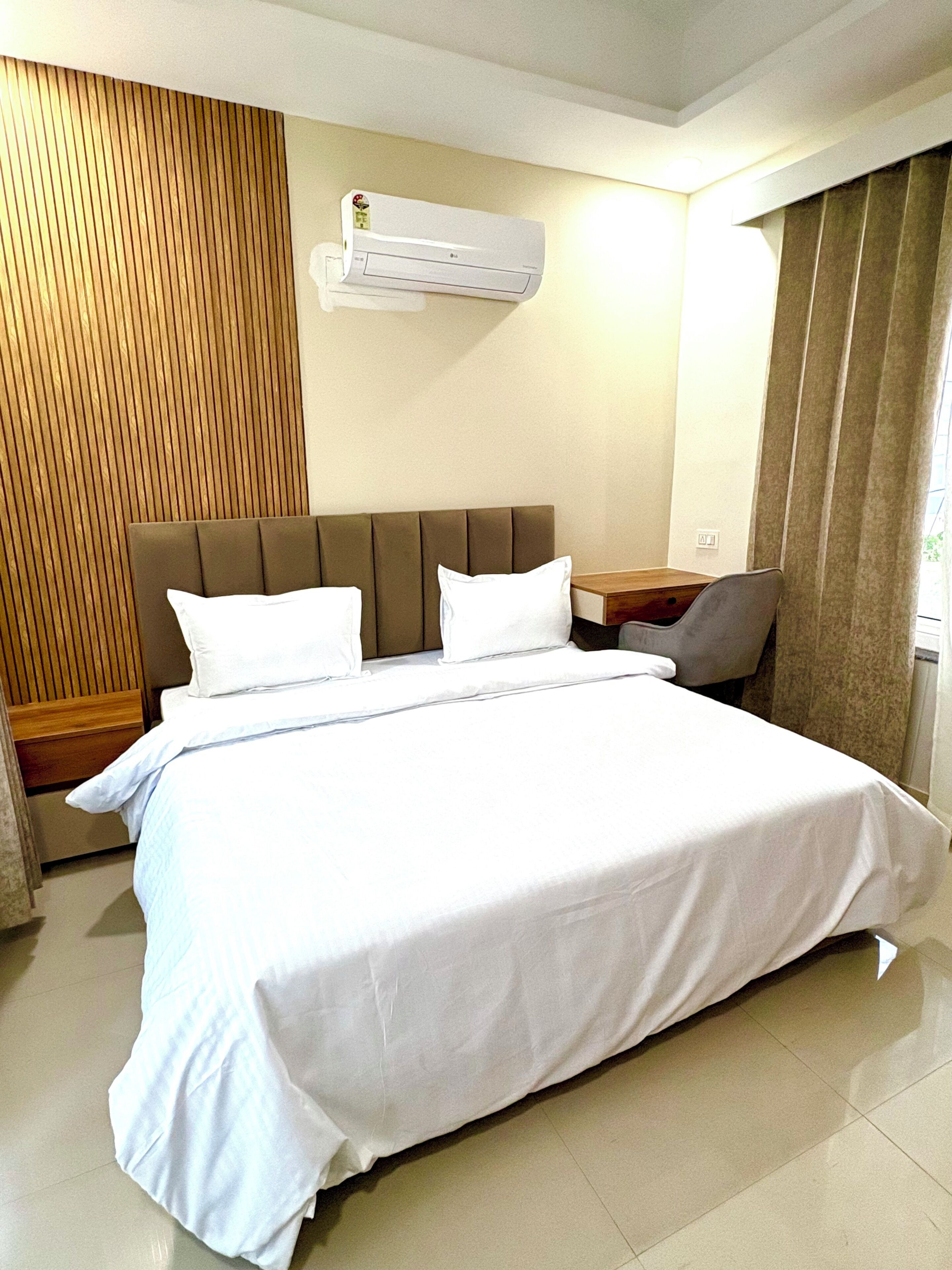 Habitación de hotel con cama, mesita de noche, unidad de aire acondicionado y ventana con cortinas.