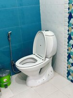 Inodoro blanco con tapa, cabezal de ducha y un cubo de basura verde en un baño con paredes de azulejos azules y blancos.