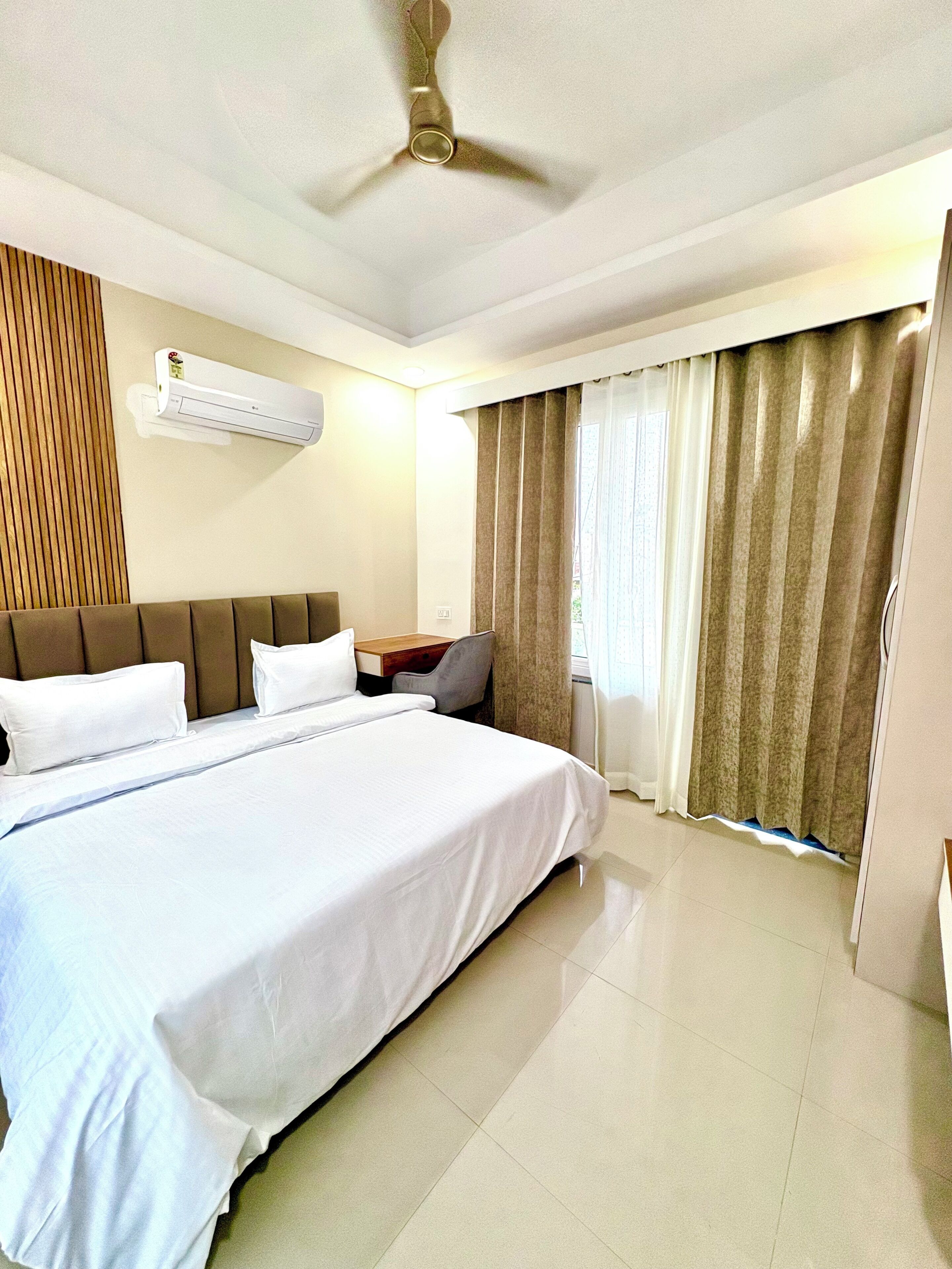 Una habitación de hotel con una cama grande, un equipo de aire acondicionado, un ventilador de techo, un escritorio con una silla y una ventana con cortinas.