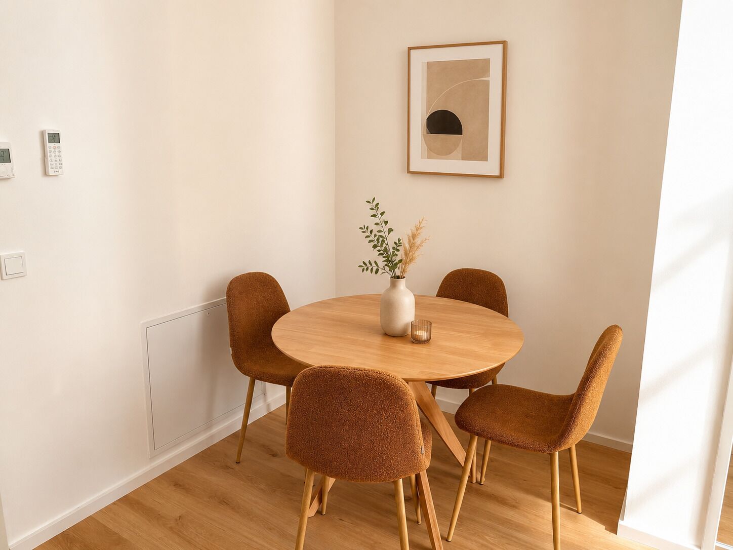 Uma sala de jantar com uma mesa redonda de madeira, quatro cadeiras castanhas, um vaso com plantas secas e uma obra de arte abstrata emoldurada na parede.