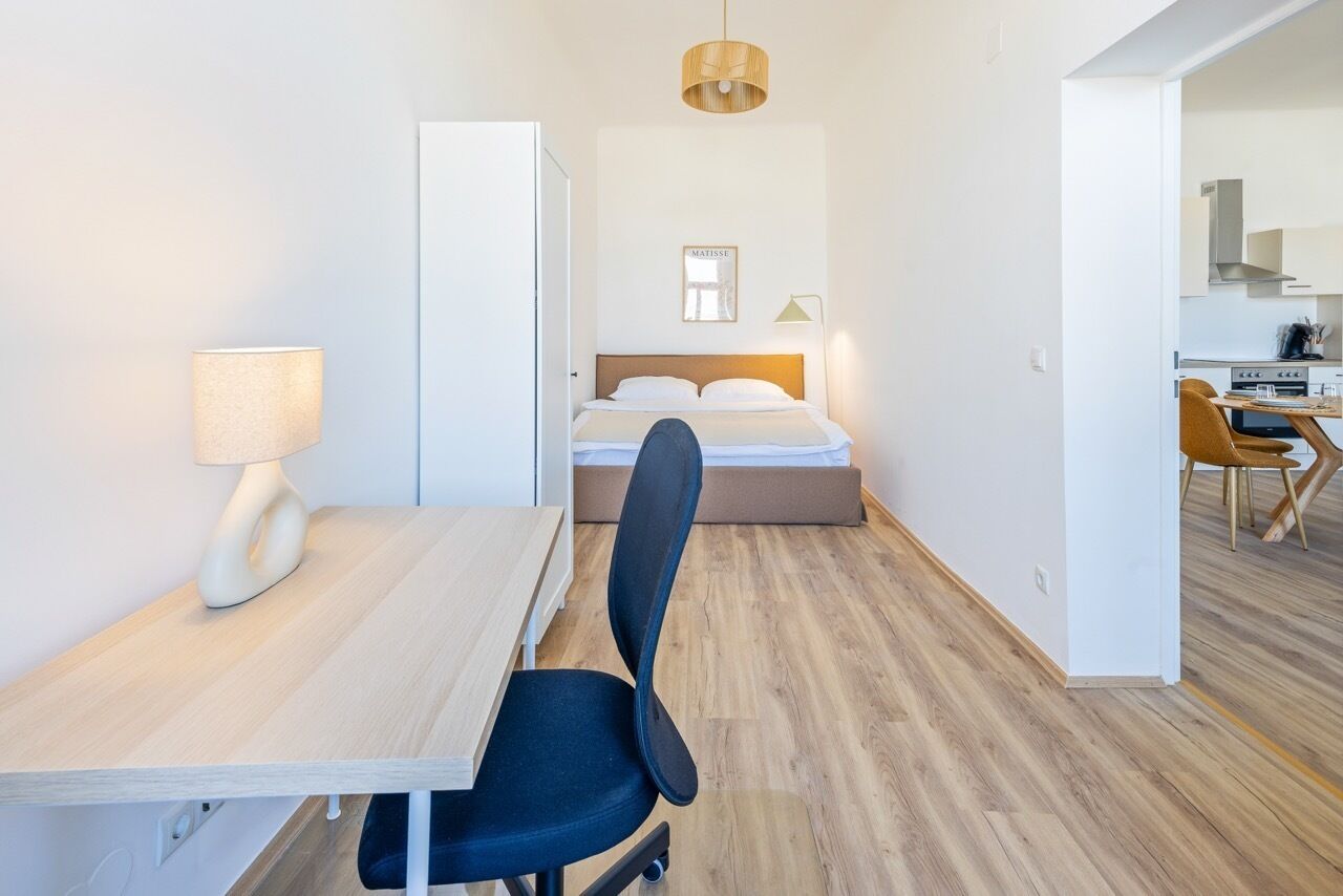 Una habitación de hotel moderna con un escritorio de madera, una silla negra, una cama con cabecera blanca y una pequeña kitchenette al fondo.