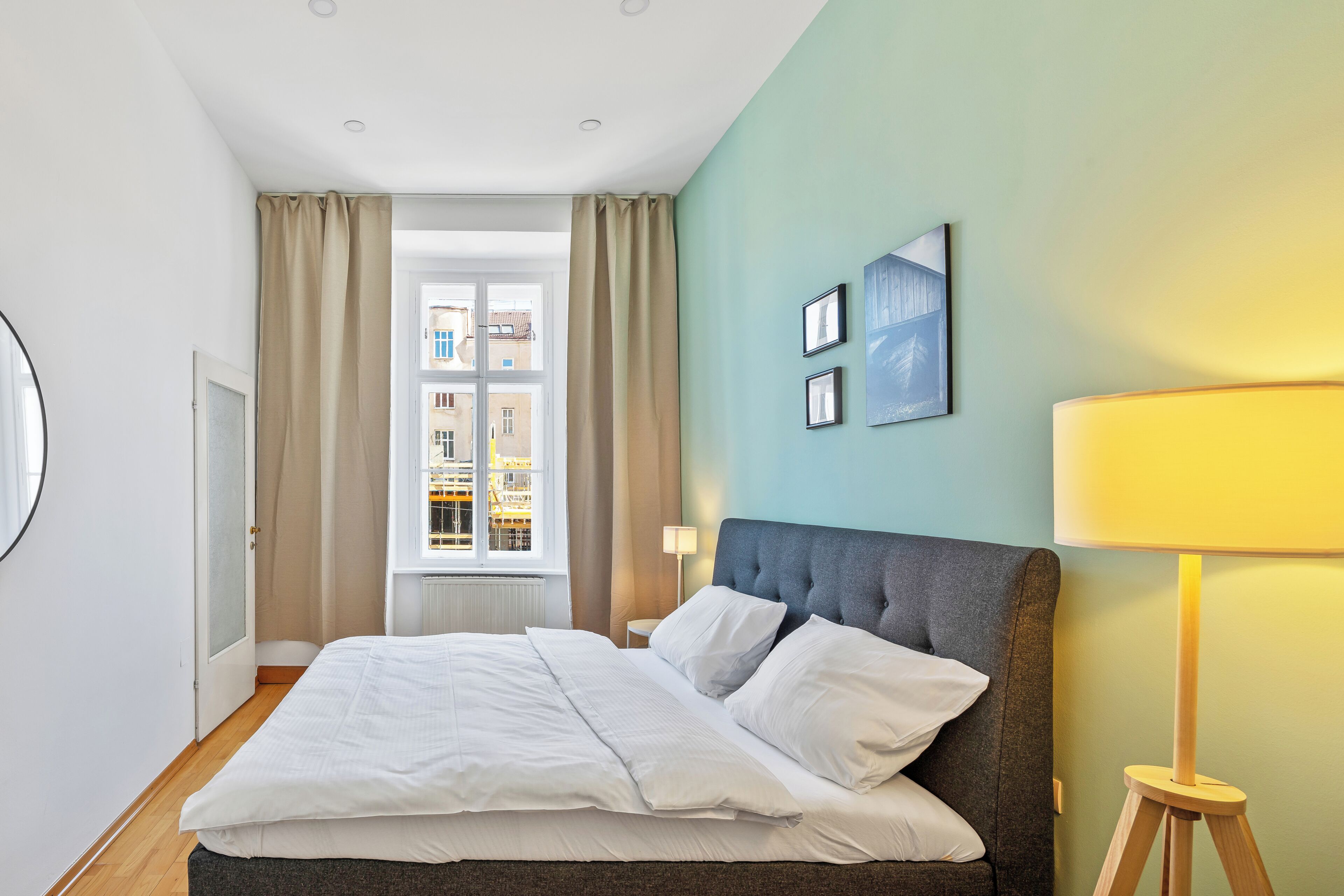 Habitación de hotel con cama, ventana con cortinas, lámpara de pared y cuadros enmarcados.