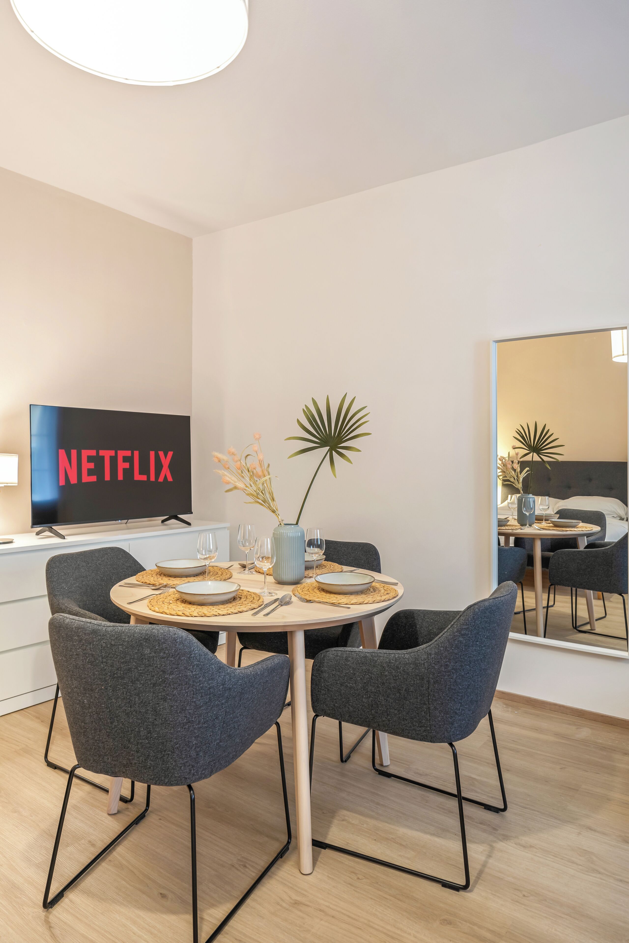 A modern dining area with a round table, four grey chairs, and a television displaying the Netflix logo.