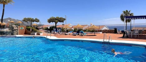 A swimming pool with clear blue water, surrounded by lounge chairs and umbrellas, with a mountain view in the background.