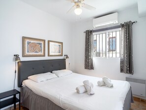 A hotel room with a bed, two framed artworks, a ceiling fan, an air conditioning unit, and a window with curtains. - Los Naranjos 11B Capistrano Village by Casasol by Interhome (Nerja)