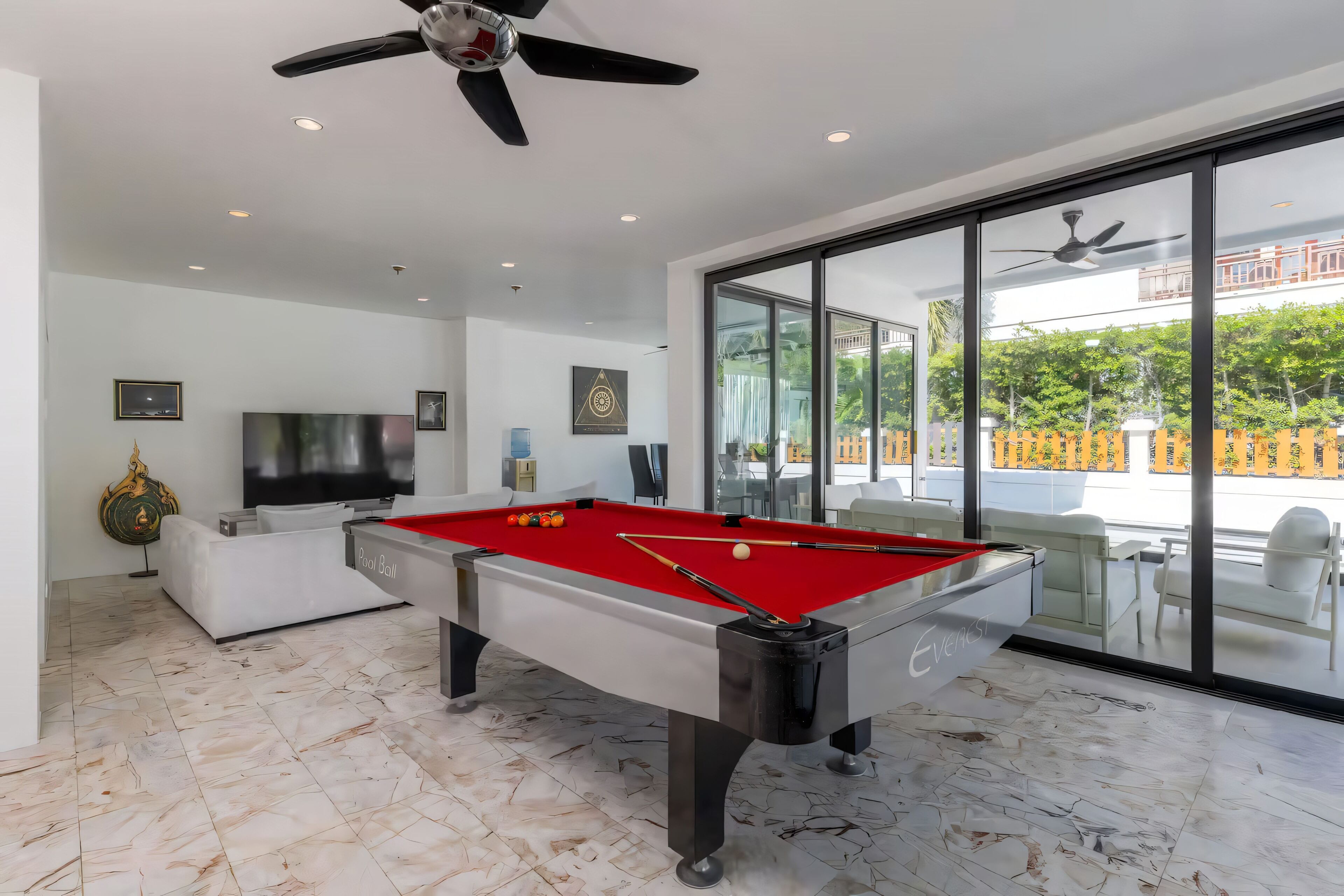 A modern indoor pool table with a red felt surface, a television, and a ceiling fan in a well-lit room with large windows.