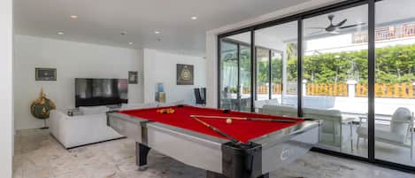 A modern indoor pool table with a red felt surface, a television, and a ceiling fan in a well-lit room with large windows.