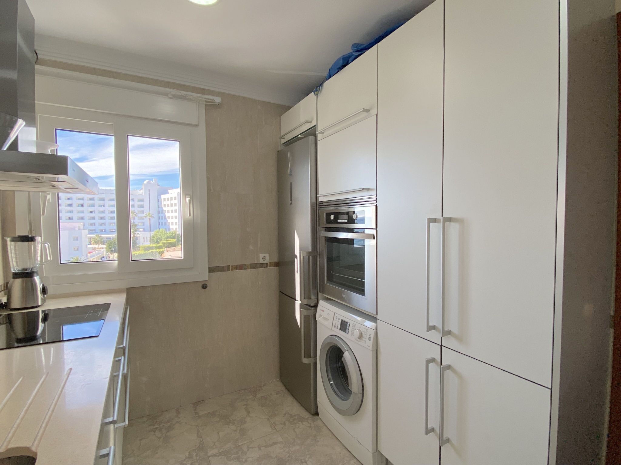 Cocina compacta con lavadora integrada, refrigerador y ventana con vistas a edificios.