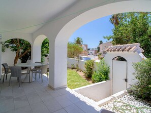 Ein überdachter Patio-Bereich mit Tisch und Stühlen, umgeben von weißen Wänden und Bögen, mit Blick auf einen Garten und eine Wohngegend.