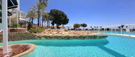 A clear blue pool with a rocky island in the center, surrounded by palm trees and a white building with columns.