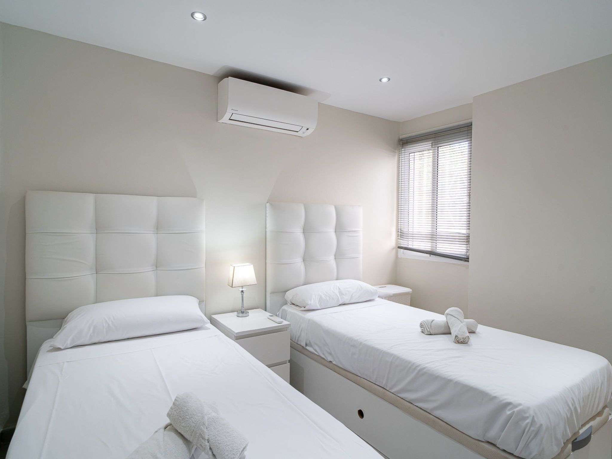 A hotel room with two beds, a bedside table with a lamp, an air conditioning unit, and a window with blinds.
