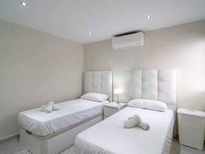 A hotel room with two beds, a bedside table with a lamp, a white air conditioning unit, and a white laundry basket. - Capuchinos 48 San Juan Capistrano by Casasol by Interhome (Nerja)