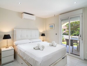 Ein Schlafzimmer mit einem großen Bett, zwei Nachttischlampen, einer Klimaanlage und Blick auf einen Außenbereich durch eine Schiebetür.