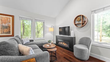 A living room with a grey sofa set, a round coffee table, a fireplace, a television mounted above it, and a window with a view of trees.