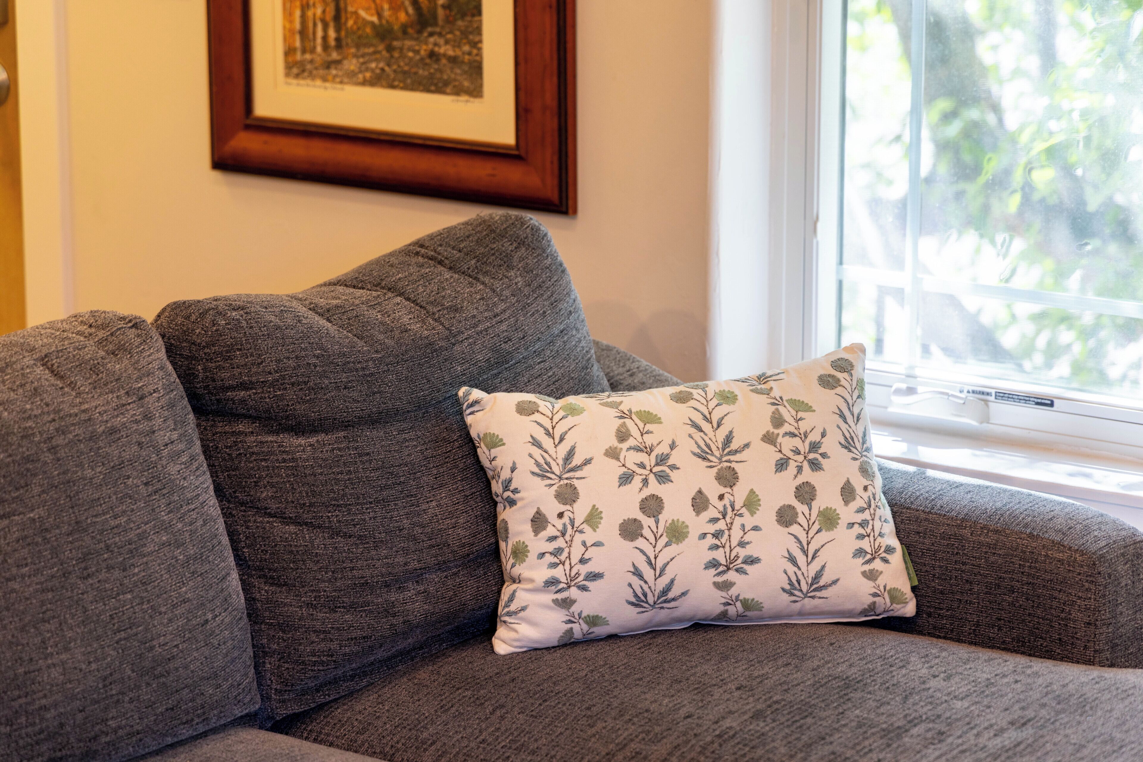 A grey sofa with a floral pillow and a framed picture on the wall.